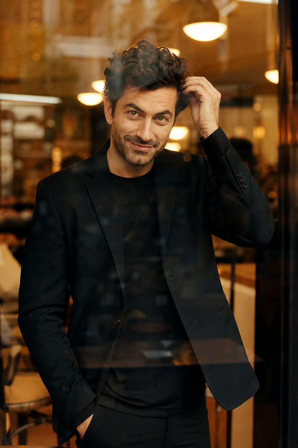 A man with thick slightly curled hair grinning as he fixes it in a cafe window reflection