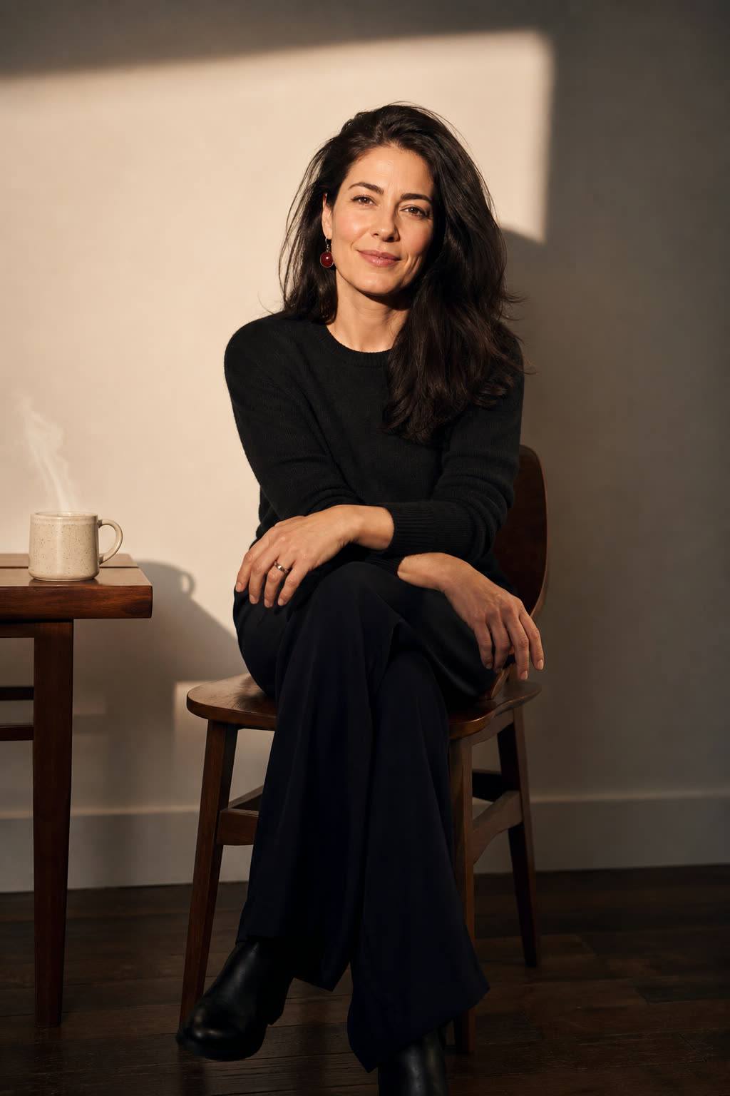 A woman in her late 30s with thick loose dark hair rim-lit against a white wall