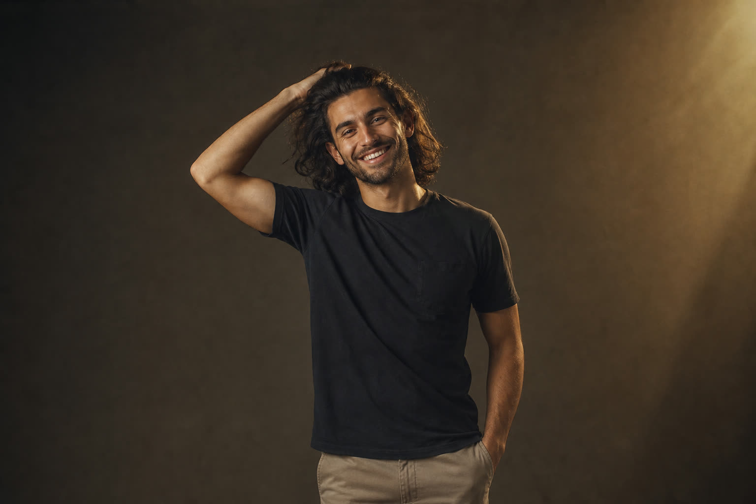 A man in his 20s with shoulder-length hair pushed back by his hand, smiling wide