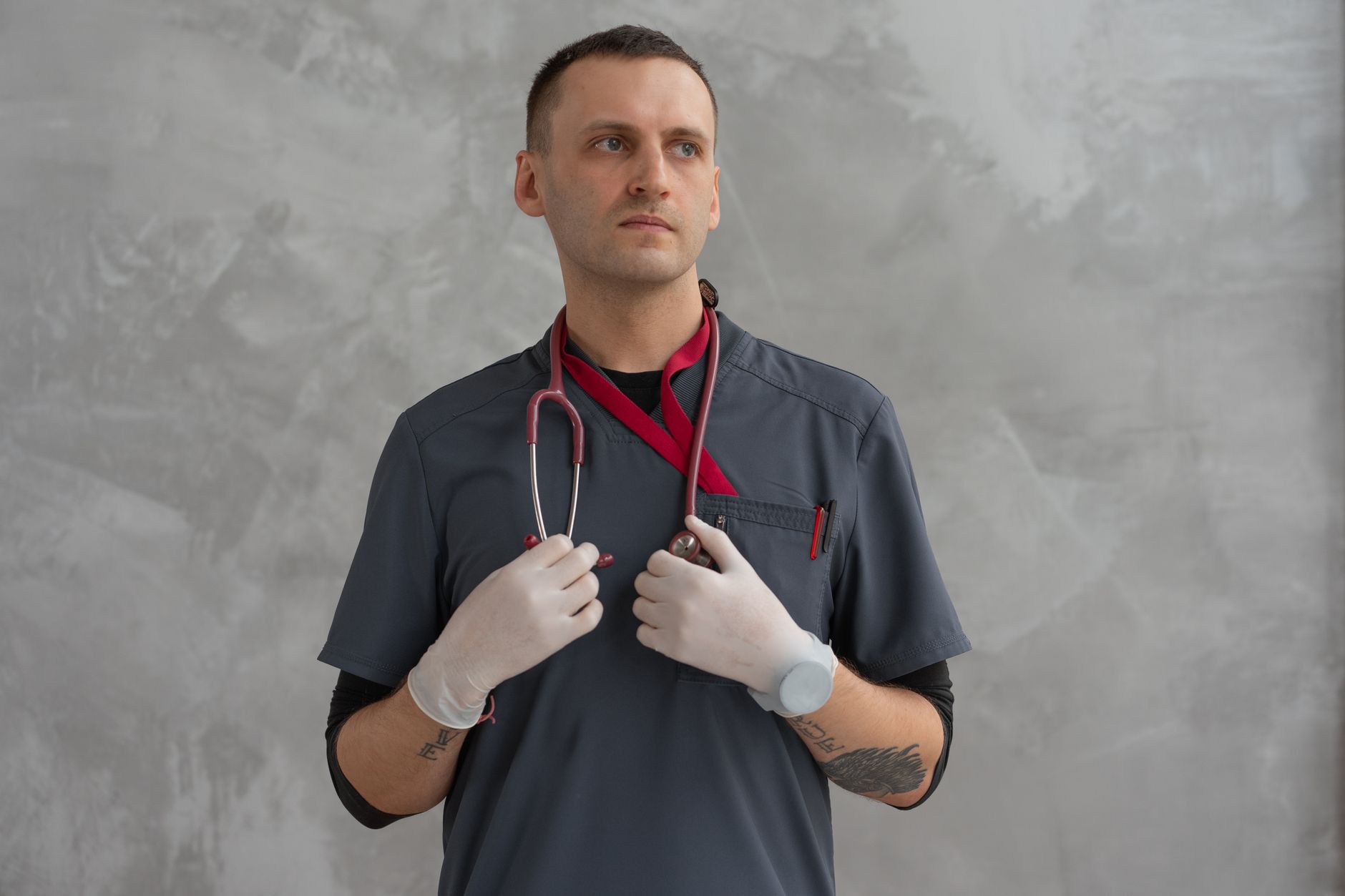 Tattooed medical professional wearing gloves and scrubs with a stethoscope on a gray background.