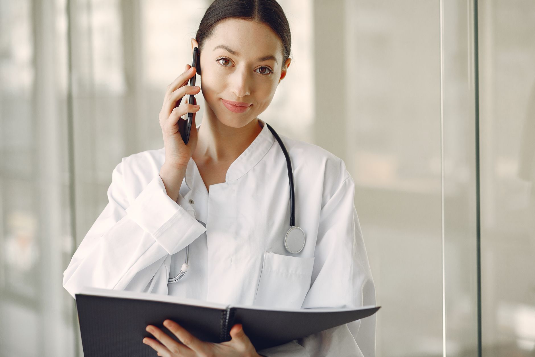 Crop ethnic female doctor in medical uniform with stethoscope and notebook talking on cellphone while standing in clinic corridor