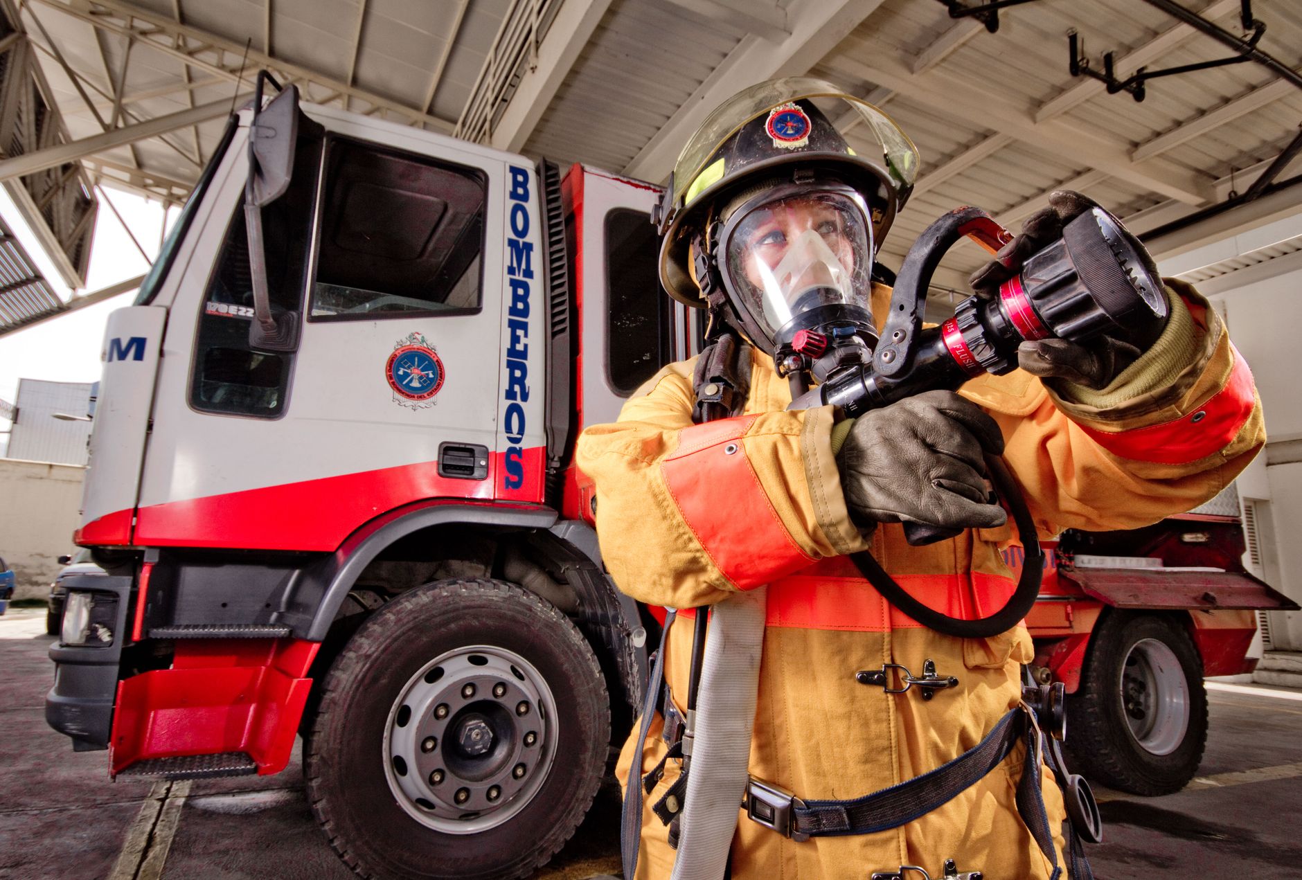 Firefighter in protective gear holding a hose near a firetruck in a garage setting.