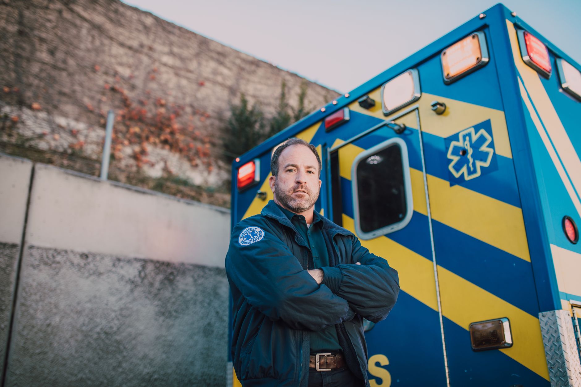 Male paramedic standing confidently beside a blue and yellow ambulance outdoors.