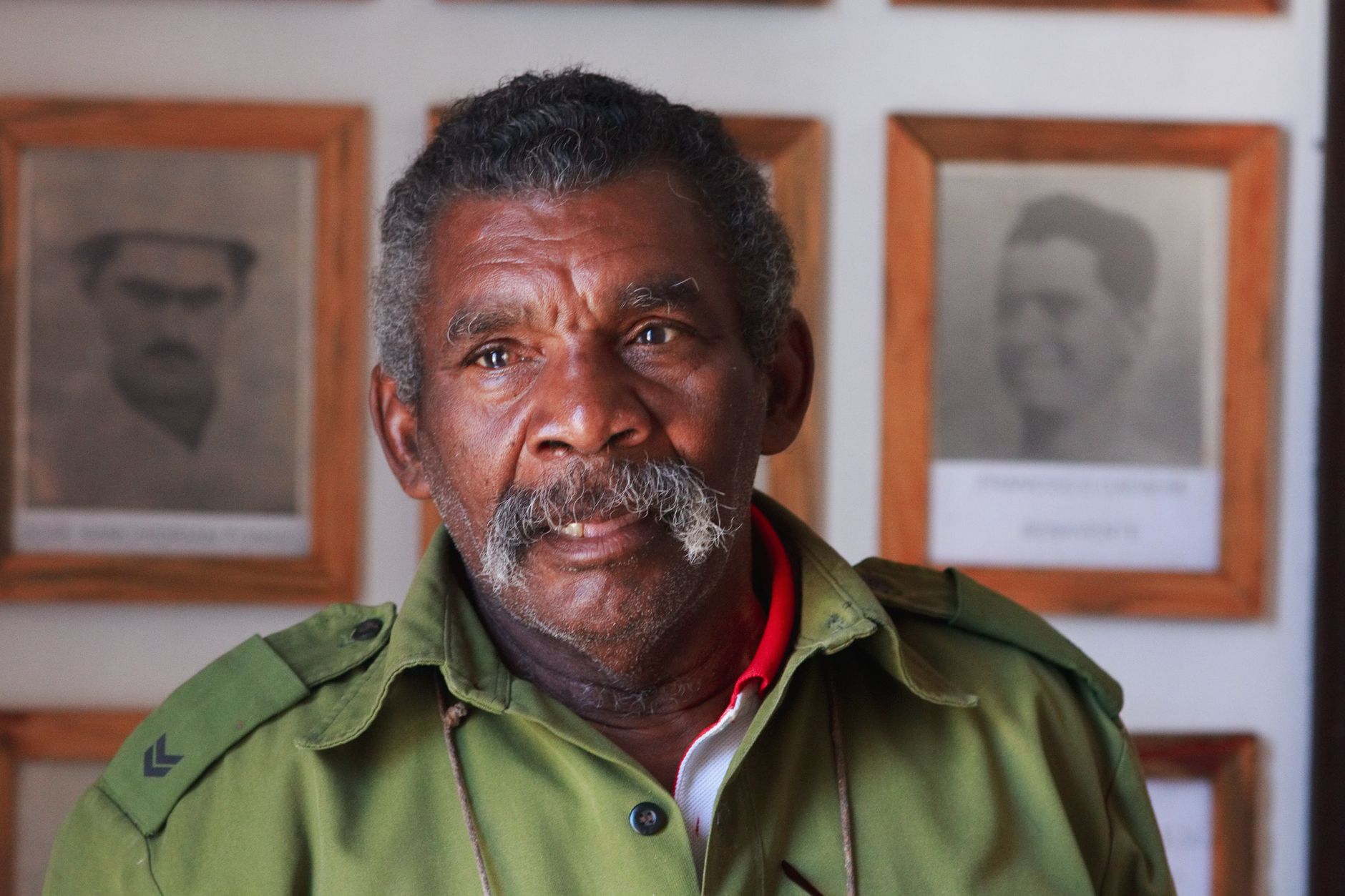 Portrait of an elderly man in uniform with a pensive look, surrounded by historical photographs.