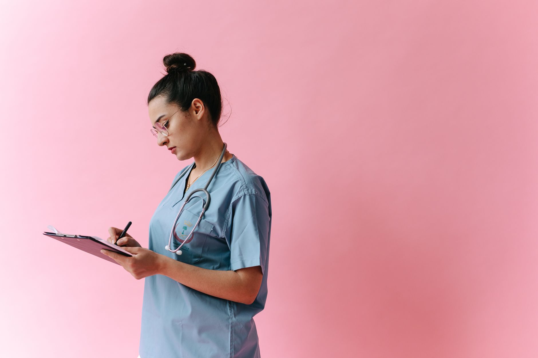 Female nurse in scrubs writing on a clipboard with a stethoscope on a pink background.