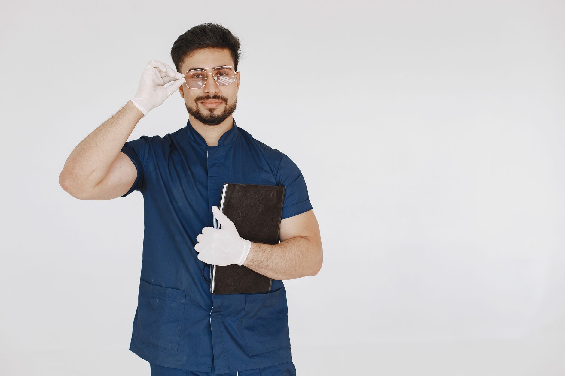 Healthcare professional wearing blue scrubs and gloves holding a clipboard indoors.