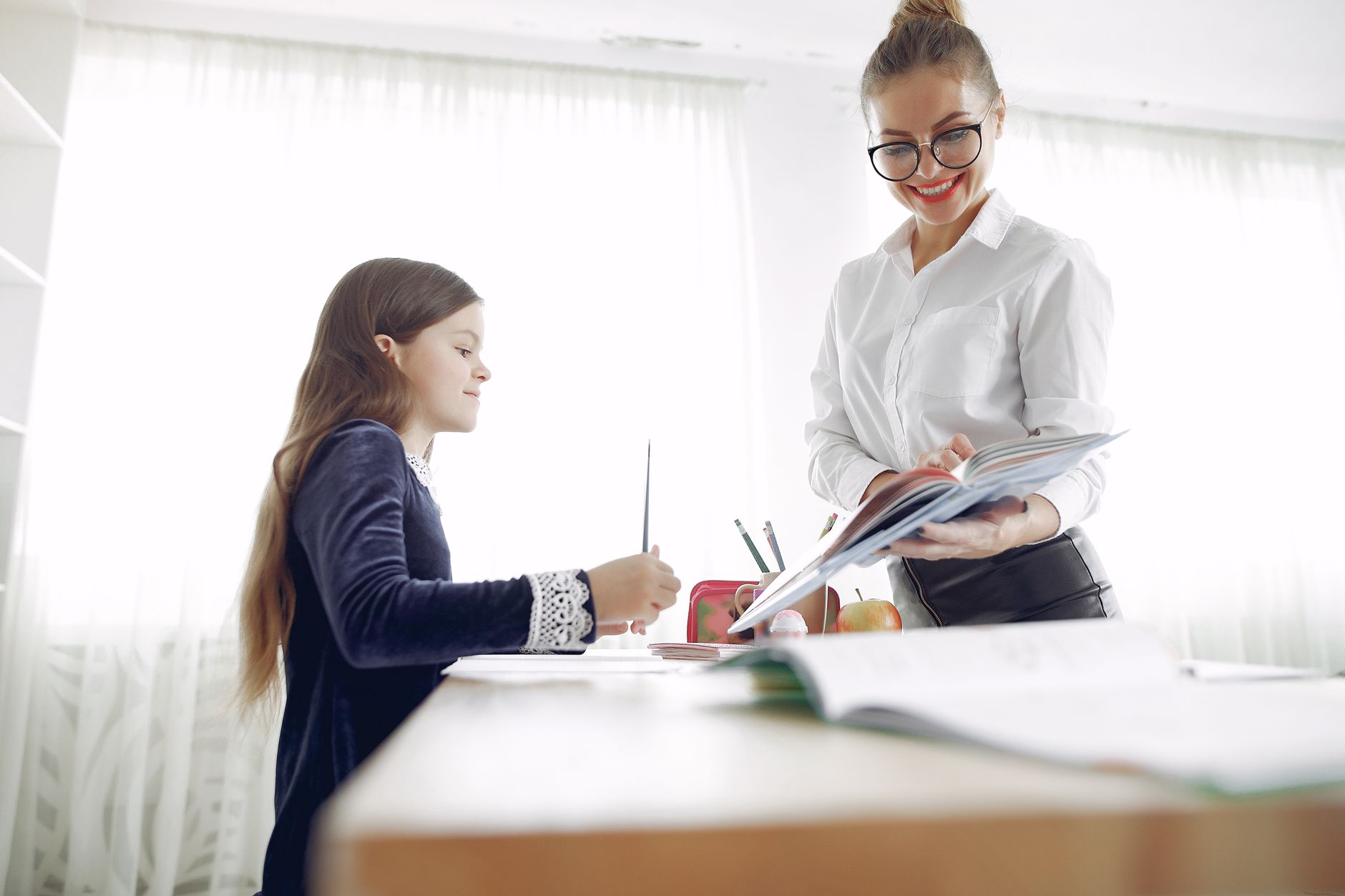From below of smiling female in formal wear and glasses tutor explaining homework task to little girl in creative place