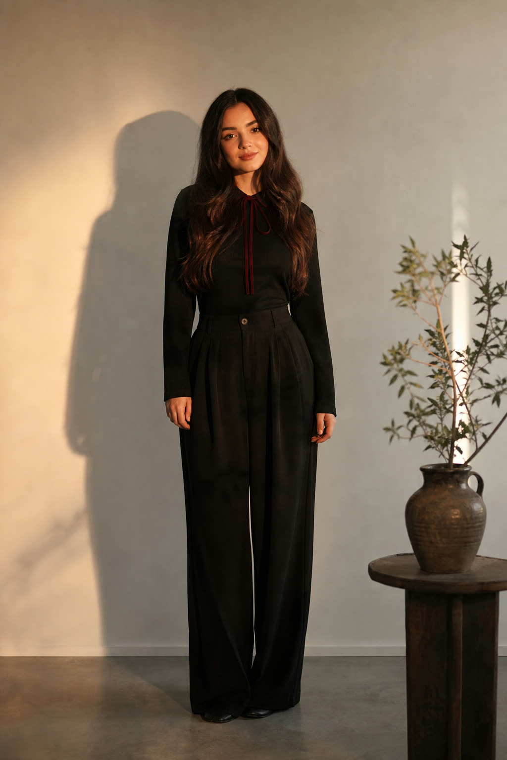 A woman in her mid 20s with thick dark hair rim-lit in warm light against a white wall
