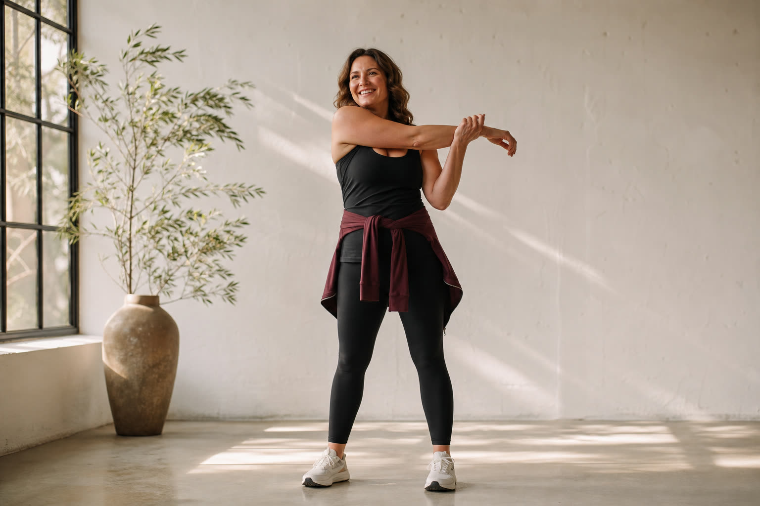 A strong woman in her 40s stretching happily in a white studio crossed by warm light and deep shadow
