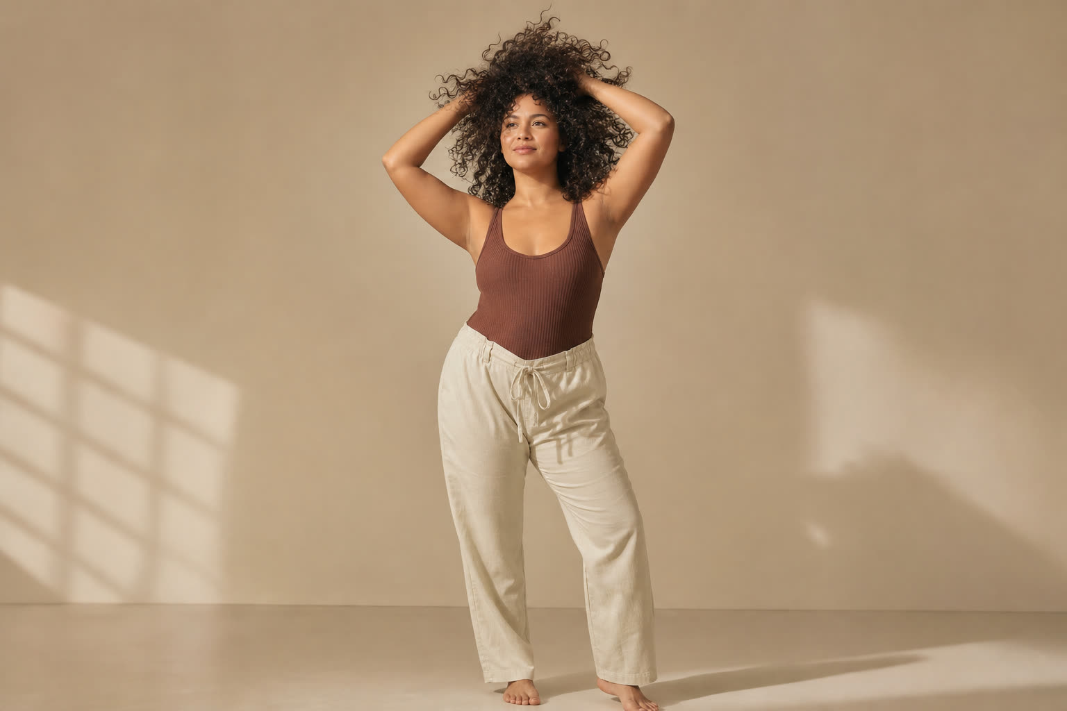 A woman in her 20s with a soft curvy build and glorious tight curls, both hands lifted into her hair