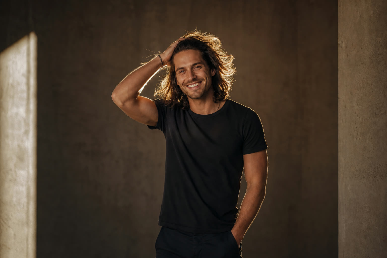 A man in his 20s with shoulder-length hair pushed back by his hand, smiling wide