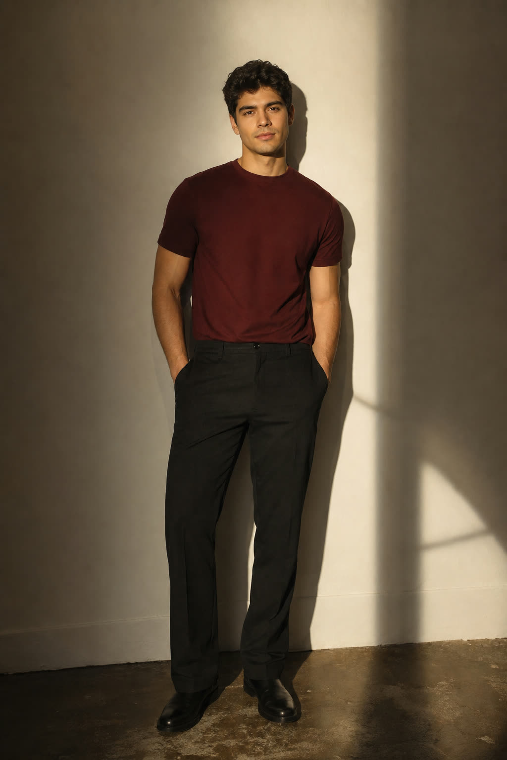 A young man with thick dark hair in an oxblood tee, lit in warm low light against a shadowed wall