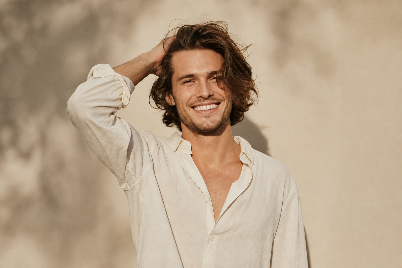 A man in his 20s with shoulder-length hair pushed back by his hand, smiling wide