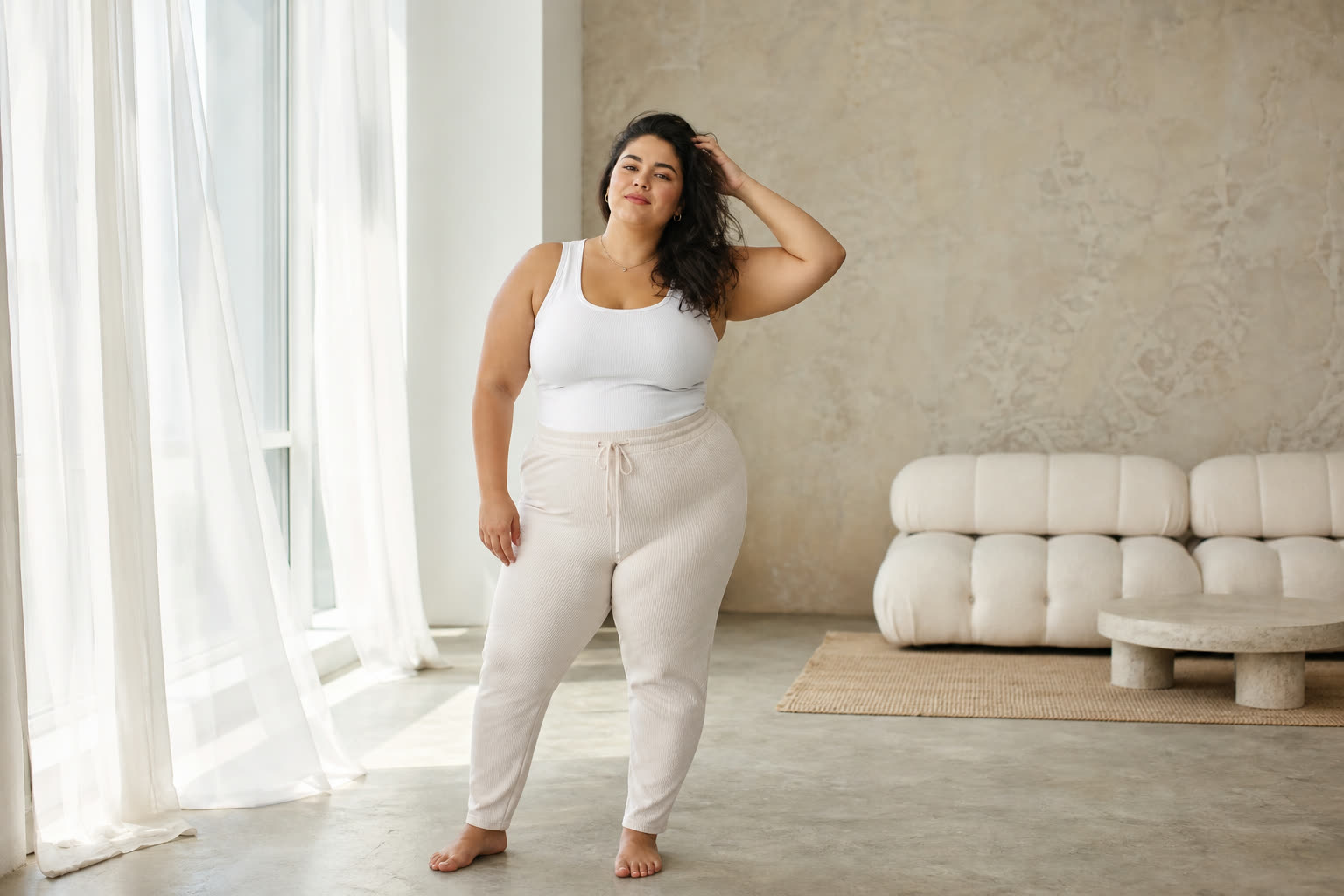 A mid-20s woman with a plus-size build running a hand slowly through thick regrown hair by a tall window
