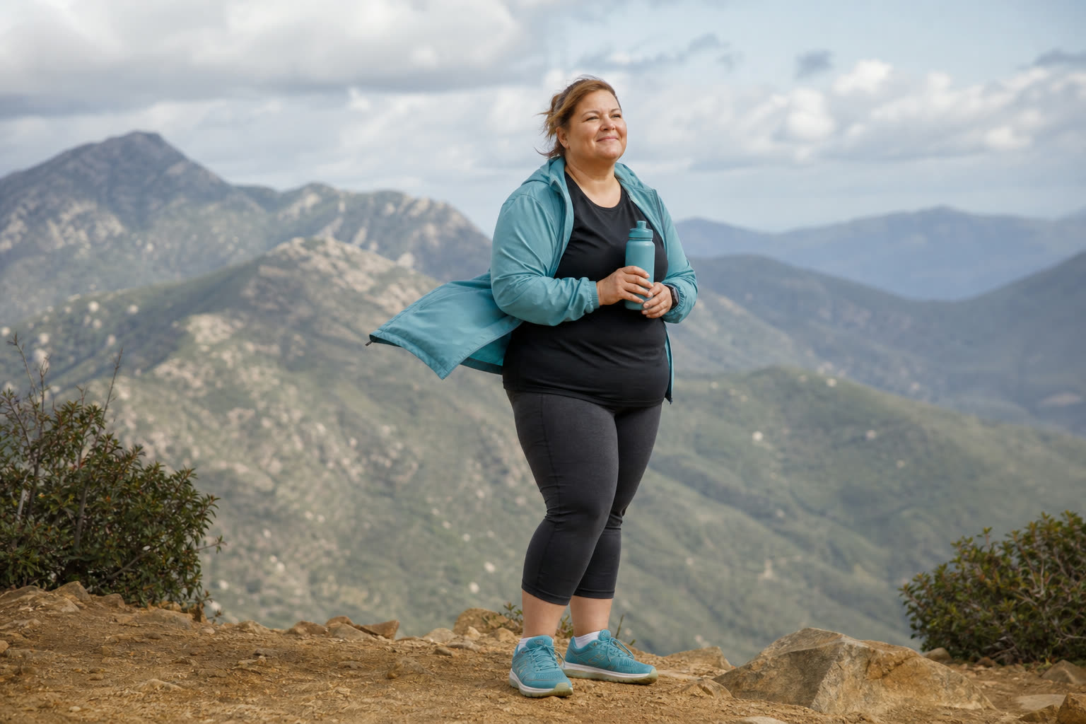 A early 50s woman with a stocky build pausing at a hiking lookout with a water bottle, wind in their clothes