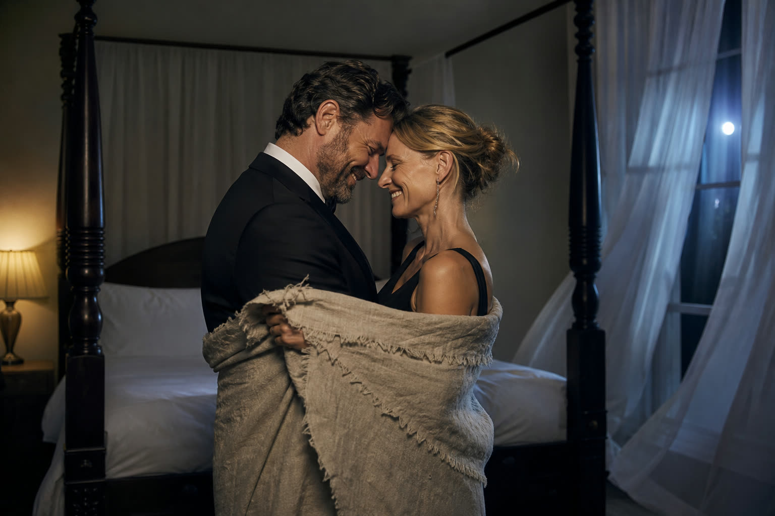 A couple standing at the foot of the bed in soft lamplight, arms around each other, smiling