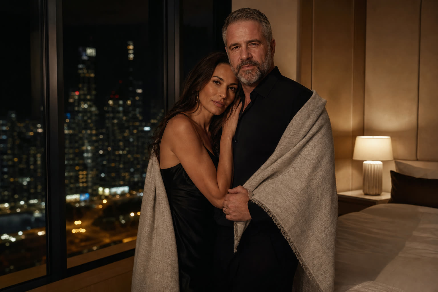 A couple standing entwined beside a tall bedroom window, city lights far below