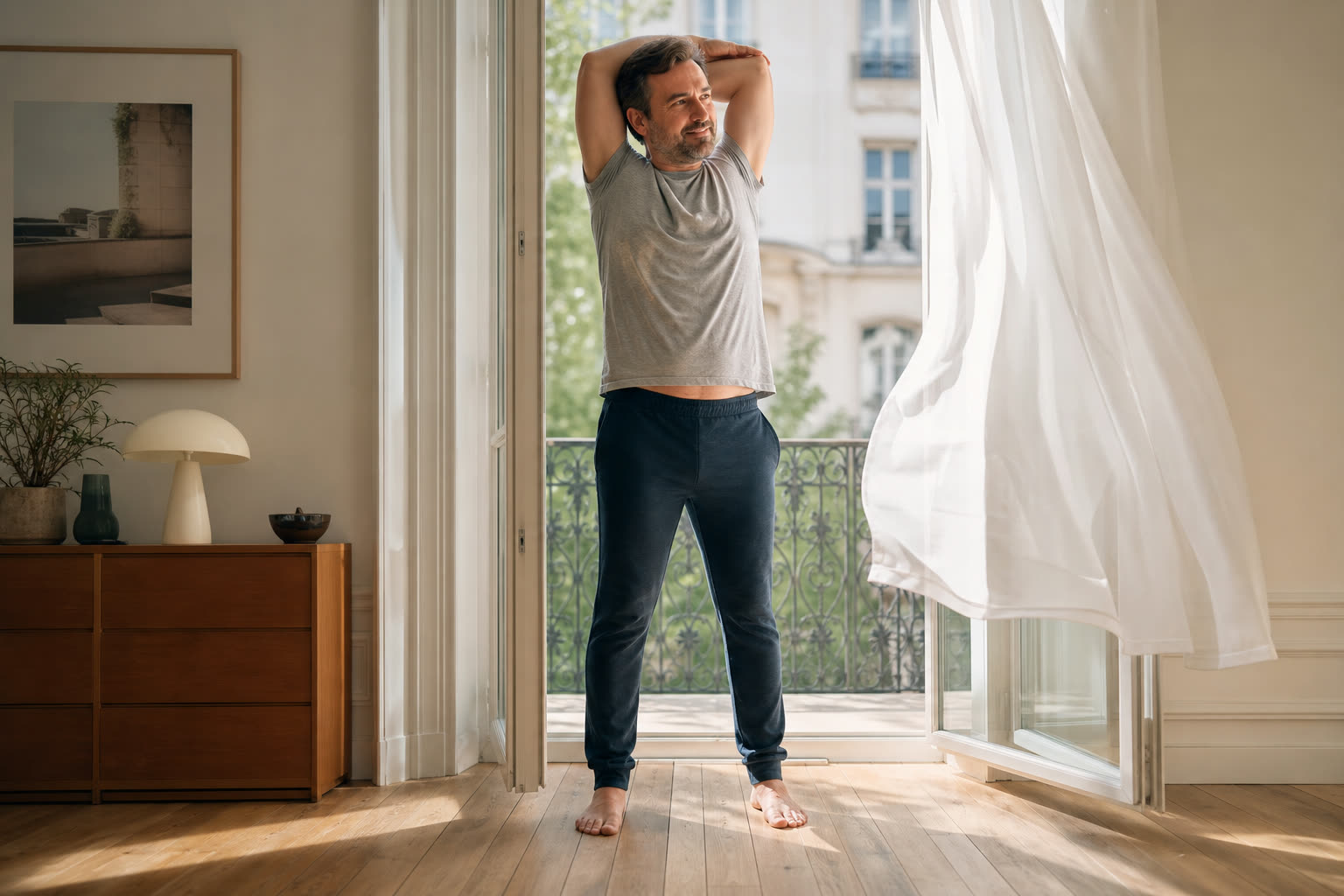 A 40s man with a soft curvy build stretching by an open balcony door in morning light, energized