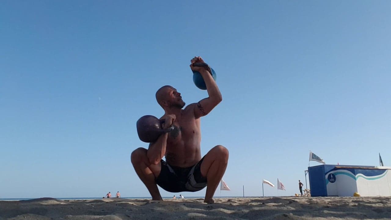 A strong man doing kettlebell squats on a beach