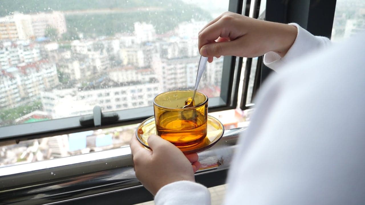 A person stirring tea by a tall window, steam rising